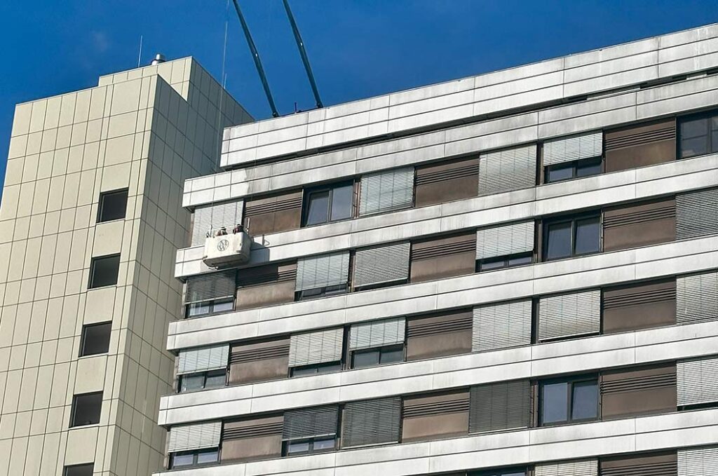 Ein Hochhaus mit grauen Fassaden und mehreren Fenstern mit heruntergelassenen Rollläden vor klarem Himmel.